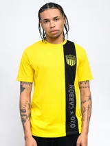Remera amarilla con una franja vertical negra en el lado izquierdo. En la parte superior de la franja se encuentra el escudo del Club Atlético Peñarol. En la parte inferior de la franja se lee la leyenda "Oro y Carbón".