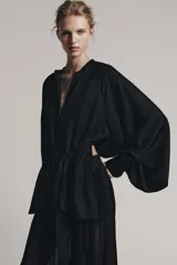 Camisa negra de cuello redondo y manga larga ancha con silueta fluida, con cierre frontal de botones y aberturas laterales en el bajo.