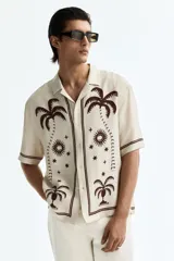Camisa de manga corta color beige, con cuello abierto y cierre frontal de botones. Presenta un estampado en marrón de palmeras, soles y estrellas, con líneas en los bordes.