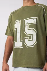 Remera de algodón con estilo deportivo, color verde oliva con efecto de lavado gastado. Presenta el número 15 estampado en blanco en el frente y detalles de costuras con vivos blancos en los hombros. Posee cuello redondo y corte holgado.