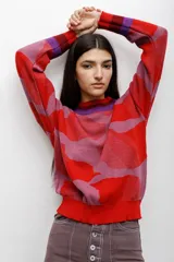 Buzo tejido en técnica jacquard con diseño de formas abstractas en tonos rojos, rosados y violetas. Presenta cuello redondo, mangas largas y puños y ruedo acanalados.
