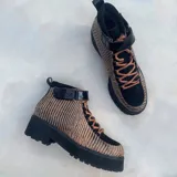 Botas estilo borceguíes en tonos beige y negro, con estampado texturizado de rayas verticales, pasadores metálicos para cordones marrones, tira con velcro en el tobillo en charol negro y suela gruesa tractorada negra.