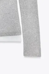 Camiseta de manga larga gris melange con cuello redondo y diseño asimétrico que simula una doble camiseta, con una capa inferior blanca visible en el cuello y el bajo.