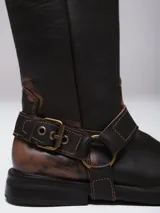 Bota de caña alta estilo western confeccionada en cuero con acabado avejentado. Presenta punta cuadrada, taco bajo y una correa con estribo metálico desmontable que rodea el tobillo.