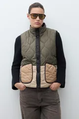 Chaleco capitoneado reversible con cuello alto, cierre frontal con botones a presión y bolsillos delanteros. Presenta un diseño bicolor en tonos verde oliva y beige con detalles en negro.
