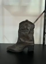 Botas texanas marrones de caña baja con detalle de tachas doradas, taco cuadrado y punta fina. Presentan un diseño estilo cowboy. Se recomienda pedir un talle más del habitual.