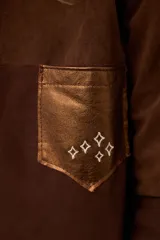 Sobrecamisa de gamuza color marrón con apliques de cuero metalizado en tono cobre en los hombros y bolsillo frontal. Presenta detalles bordados en las mangas, el bolsillo y la espalda, con un diseño de corte recto y cuello camisero.