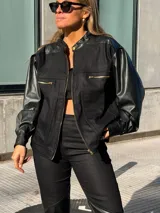 Campera de estilo bomber combinada en denim negro y mangas de cuero sintético, con cierre frontal y bolsillos con cremallera en el pecho.