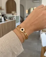 Esclava rígida de acero con baño de oro, diseño calado con detalle circular central en nácar y ónix, decorada con dos circonias. Incluye una cadena fina adicional integrada.