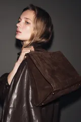 Bolso de mano formato city confeccionado en gamuza color marrón oscuro. Cuenta con doble asa de mano, asa bandolera extraíble, compartimento interior con cierre y bolsillo exterior con imán.