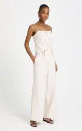 Pantalón sastrero de lino y algodón color beige, de tiro alto y corte ancho (wide leg). Presenta sutiles pliegues frontales, trabillas con apliques metálicos dorados en la cintura y bolsillos laterales.