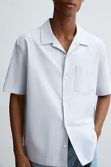 Camisa de algodón con rayas verticales finas azules y blancas, corte relajado (relaxed fit), cuello bowling y manga corta. Posee un bolsillo plastrón en el pecho y aberturas laterales en el bajo.
