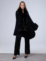 Ruana tejida de corte amplio y diseño elegante, con cuello, bordes frontales y puños adornados con apliques de piel sintética negra. Presenta una abertura central con cierre de botón tipo trenca.