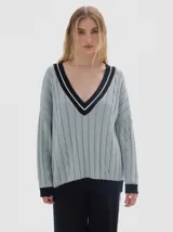 Buzo tejido de punto con diseño oversized, cuello en V profundo y mangas largas. Presenta un patrón de trenzas verticales en color verde oscuro con detalles en contraste de color rosado en el cuello, puños y ruedo.