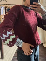 Sweater de punto color bordó con cuello redondo y mangas largas. Presenta un diseño de guardas geométricas en zigzag en tonos gris y verde claro en la parte inferior de las mangas.