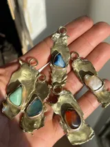 Collar artesanal con dije de base metálica irregular y piedra central roja. Presenta una estructura de tiento de gamuza marrón con detalles de tubos metálicos dorados.