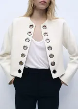 Chaqueta de corte recto y diseño estructurado en color crudo, con cuello redondo y manga larga. Presenta una abertura frontal sin cierre, decorada con ojales metálicos plateados a lo largo de los bordes. Confeccionada en tejido mezcla de algodón con forro interior.