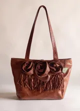 Cartera shopper de cuero color marrón terra, con doble asa de hombro y diseño decorativo de tres flores de cuero y flecos en el frente.