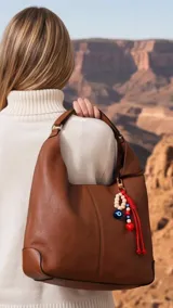 Cartera tipo hobo de cuero sintético color suela, con interior espacioso y costuras decorativas frontales. Incluye asa de hombro fija, monedero rectangular colgante a juego y un llavero amuleto con cuentas de madera, ojos turcos y detalles en rojo.