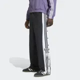 Pantalon deportivo Adidas modelo Adibreak, de corte recto y color negro. Presenta las icónicas tres tiras blancas laterales y botones a presión a lo largo de las piernas, con logo de la marca bordado en la parte inferior.