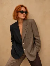 Blazer oversize de diseño bicolor con estructura asimétrica, combinando un lado en color negro y otro en color marrón, ambos con estampado de rayas diplomáticas. Presenta solapas clásicas, cierre frontal con botones de strass y un pin decorativo en forma de perro salchicha.