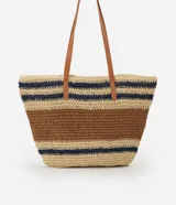 Bolso tipo tote de paja tejida con diseño de rayas horizontales en color natural, marrón y azul marino. Posee asas de hombro en cuero sintético color camel y un dije metálico circular en el frente.
