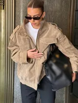Chaqueta de gamuza en color marrón oscuro, con cuello alto, cierre frontal oculto y bolsillos tipo cargo en el pecho. Presenta un diseño de corte holgado con detalles de charreteras en los hombros.