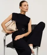 Mono negro sin mangas, con cuello redondo y cierre tipo gota en la espalda. Presenta hombreras marcadas, cintura ajustable con cinturón de lazo y hebilla metálica irregular, y pantalón ancho de pierna acampanada.