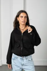 Campera de gamuza negra con cierre frontal, cuello camisero y dos bolsillos frontales con solapa. Presenta puños con broche a presión y ruedo elástico.