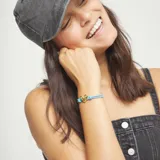 Pulsera ajustable con doble hilo elástico de color celeste brillante, centrada por un dije de corazón tridimensional con acabado dorado. Presenta un cierre o ajuste de forma ovalada dorada en la parte superior y un nudo decorativo a cada lado del corazón.