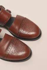 Zapato tipo monk strap con aberturas laterales, confeccionado en cuero vacuno metalizado color bronce. Presenta detalles de perforaciones estilo brogue en la puntera y el talón, y doble hebilla metálica en el empeine.