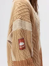 Cardigan oversize de tejido de punto grueso con diseño de bloques de color en tonos tierra. Presenta cuello en V, cierre frontal con botones, mangas largas con terminación abullonada y parches decorativos en el pecho y hombro.