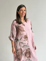 Vestido largo color rosa, con mangas cortas y diseño holgado, presenta un estampado floral en tonos marrones y verdes en un lateral, además de un bolsillo aplicado en la parte frontal y costuras visibles de estilo rústico.