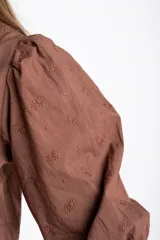 Blusa blanca de algodón con delicado bordado floral, mangas abullonadas de largo 3/4 y cierre frontal con botones y cordón ajustable en la cintura.