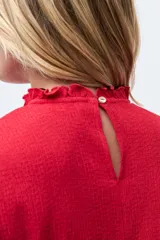 Blusa de color rojo con cuello alto y detalle de volados, mangas largas con puños elásticos en punto smock y textura sutil en la tela.