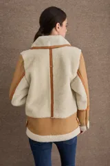 Campera tipo gamulán de cuero color camel con interior y detalles de piel sintética en color crema. Presenta cuello alto con doble hebilla ajustable, cierre frontal con cremallera y bolsillos laterales con cierre.