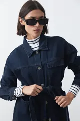 Sobrecamisa de jean color negro con silueta relajada, cuello camisero, cierre frontal con botones metálicos, bolsillos frontales y cinturón ajustable en la cintura.