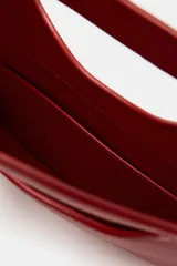 Bolso de mano de piel color rojo, con diseño de cuerpo asimétrico y asa de mano integrada con apertura lateral. Incluye correa bandolera extraíble y bolsillo interior.