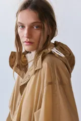 Cazadora tipo anorak color beige, con capucha ajustable, manga larga y bolsillo frontal de plastrón con solapa. Cuenta con cordones elásticos ajustables en la cintura y el bajo, y cierre frontal con botones a presión.