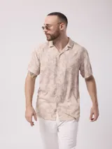 Camisa de manga corta color beige con estampado de hojas en tonos marrones. Confeccionada en viscosa, tiene cuello tipo solapa y cierre frontal de botones.