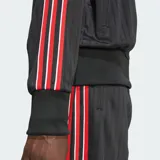 Campera deportiva Adidas modelo Firebird Loose Pinstripes, de color negro con diseño de rayas diplomáticas sutiles. Cuenta con cuello alto, cierre frontal completo, bolsillos laterales y bandas tricolor en rojo, blanco y negro a lo largo de las mangas. Incluye el logo del trifolio bordado en el pecho.