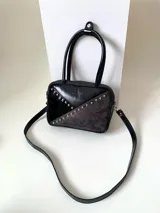 Cartera rígida negra con detalles en rojo y tachas plateadas. Tiene herrajes y cierre metálico plateado marca YKK, asa fina para llevar al hombro o en la mano y correa larga extra regulable. El interior es forrado, tiene un bolsillo con cierre enmarcado en cuero, y un segundo bolsillo plaqué.