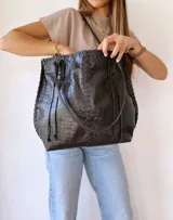 Cartera tipo tote de cuero negro con textura de cocodrilo. Presenta detalles de costuras trenzadas a lo largo de la estructura y tiras decorativas laterales. Cuenta con doble asa de hombro y cierre superior.