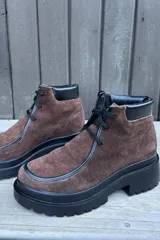 Botas de caña corta confeccionadas en cuero nobuk color marrón, con detalles en cuero negro en los bordes y cordones negros. Presentan una suela de plataforma dentada de color negro.