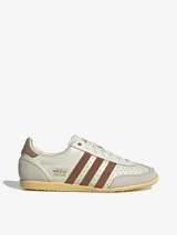 Championes Adidas modelo Japan, con diseño retro de perfil bajo. Presentan un cuerpo de cuero color crema con perforaciones en la puntera, icónicas tres tiras laterales en color marrón y refuerzos de gamuza gris en la puntera y el talón. Incluye suela de goma color amarillento y detalles dorados en el lateral y la lengüeta.