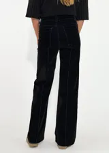 Pantalon de corte recto y tiro medio, confeccionado en denim con acabado aterciopelado en tono marrón oscuro. Presenta costuras verticales marcadas en contraste y cierre frontal con botón y cremallera.