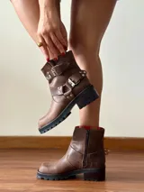 Botas cortas de cuero color caoba, con diseño de estilo biker. Presentan doble correa con hebillas metálicas y tachas decorativas en la caña, cierre lateral interno y suela track de goma negra con taco cuadrado.