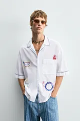 Camisa blanca de algodón con cuello bowling y manga corta, corte relajado. Presenta bordados a contraste en azul, rojo y amarillo que simulan elementos de cocina y artesanía, además de ribetes de pespunte azul en cuello, puños y abertura frontal.