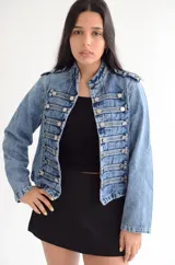 Chaqueta de estilo militar confeccionada en denim negro con efecto lavado. Presenta un diseño estructurado con cierre frontal de cremallera, cuello mao y múltiples botones metálicos decorativos dispuestos en filas horizontales. Incluye detalles de charreteras en los hombros y puños con botones.