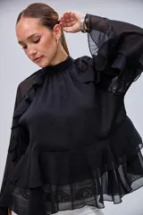 Blusa negra de cuello alto con volados en el pecho y mangas largas con transparencias. Presenta un diseño escalonado con volados en la parte inferior.